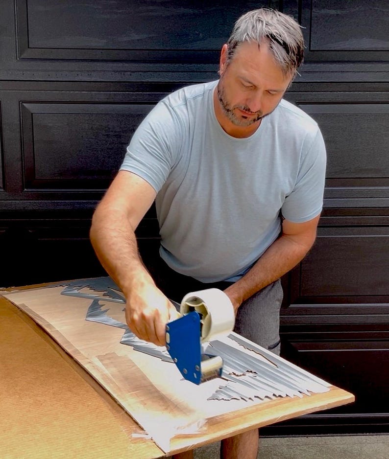 May include: A blue tape dispenser with a clear roll of adhesive tape being used to secure a silver-colored, jagged-edged metal mountain silhouette art piece onto a flat, light-brown wooden board surface for packaging and protection.