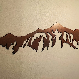 May include: A brown metal mountain range wall art sculpture. The sculpture is cut out in the shape of a mountain range with peaks and valleys.