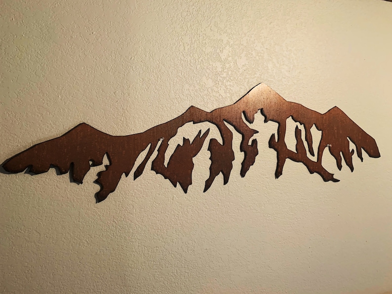 May include: A brown metal mountain range wall art sculpture. The sculpture is cut out in the shape of a mountain range with peaks and valleys.