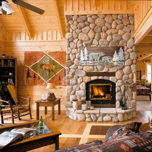 K&ouml;nnte beinhalten: Ein gem&uuml;tliches Wohnzimmer mit einem Steinkamin, Holzb&ouml;den und einer hohen, schr&auml;gen Decke. &Uuml;ber dem Kamin h&auml;ngt ein Metallkunstwerk mit B&auml;ren und B&auml;umen. Ein Gem&auml;lde eines Hirsches befindet sich an der Wand. Ein Schaukelstuhl und ein Couchtisch vervollst&auml;ndigen die rustikale Einrichtung.