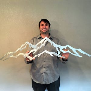 May include: A person holds a metal wall art sculpture of three white mountain peaks. The sculpture is made of thin metal and has a jagged, abstract design.