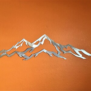 May include: A silver metal mountain range wall art sculpture with a detailed design. The sculpture is mounted on a solid orange wall.
