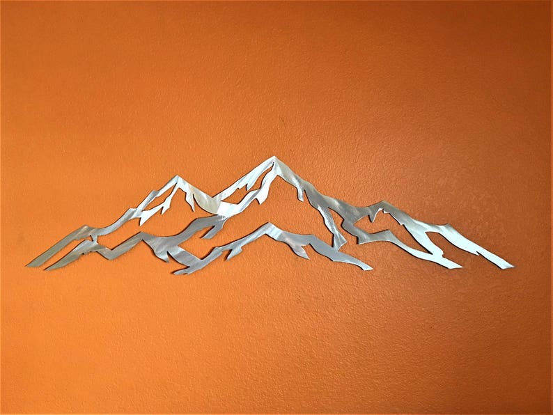 May include: A silver metal mountain range wall art sculpture with a detailed design. The sculpture is mounted on a solid orange wall.