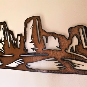 Puede incluir: Arte de pared de metal que representa un paisaje des&eacute;rtico con formaciones rocosas en capas. La obra de arte est&aacute; cortada de metal marr&oacute;n con contornos negros, creando un efecto de silueta. El fondo es de color blanco roto neutro.