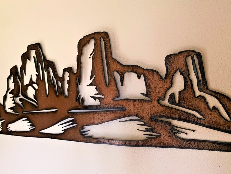 Puede incluir: Arte de pared de metal que representa un paisaje des&eacute;rtico con formaciones rocosas en capas. La obra de arte est&aacute; cortada de metal marr&oacute;n con contornos negros, creando un efecto de silueta. El fondo es de color blanco roto neutro.