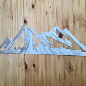 May include: A metal mountain range wall art piece with a brushed silver finish. The artwork is mounted on a wooden wall with vertical planks. The mountain range features multiple peaks and valleys, creating a detailed silhouette.