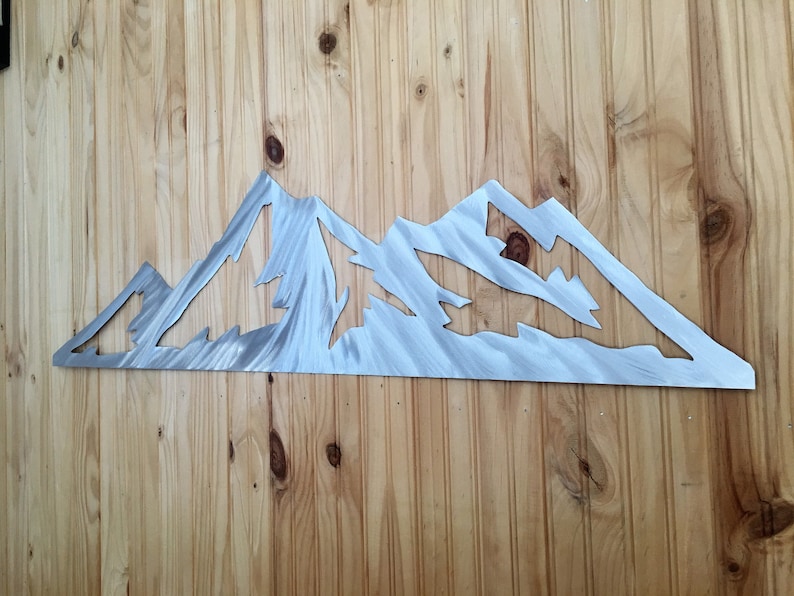 May include: A metal mountain range wall art piece with a brushed silver finish. The artwork is mounted on a wooden wall with vertical planks. The mountain range features multiple peaks and valleys, creating a detailed silhouette.