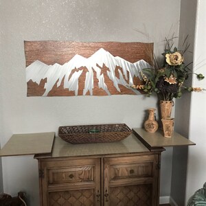 May include: A metal wall art piece depicting a mountain range with a copper-colored background and silver-colored peaks. The artwork is mounted on a light gray wall above a wooden cabinet with a woven basket and decorative vases.