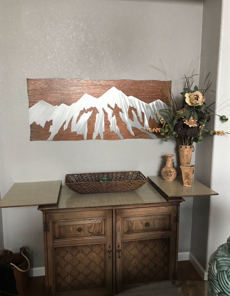 May include: A metal wall art piece depicting a mountain range with a copper-colored background and silver-colored peaks. The artwork is mounted on a light gray wall above a wooden cabinet with a woven basket and decorative vases.