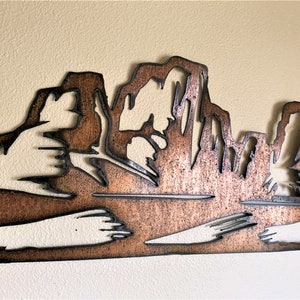 Puede incluir: Una pieza de arte mural de metal color cobre que representa una silueta de cordillera. El dise&ntilde;o presenta formas recortadas, creando un efecto de capas. La obra de arte est&aacute; montada en una pared color crema, agregando contraste.