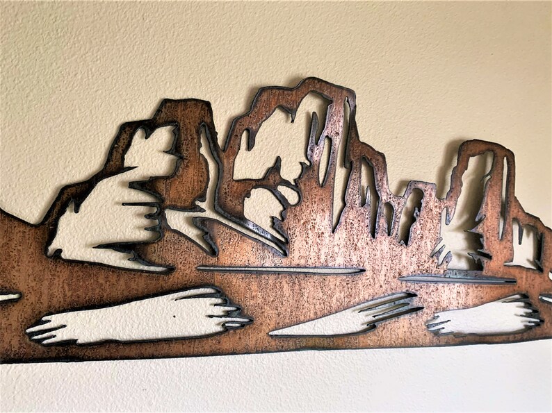 Puede incluir: Una pieza de arte mural de metal color cobre que representa una silueta de cordillera. El dise&ntilde;o presenta formas recortadas, creando un efecto de capas. La obra de arte est&aacute; montada en una pared color crema, agregando contraste.