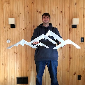 May include: A white, multi-layered mountain range wall art piece featuring jagged, snow-capped peaks. The sculpture consists of several overlapping, angular mountain silhouettes arranged in a horizontal formation, creating a layered effect.