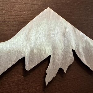 K&ouml;nnte beinhalten: Ein silberfarbener Metallzuschnitt in Form eines Gebirgszuges mit geb&uuml;rsteter Oberfl&auml;che. Das St&uuml;ck weist scharfe, gezackte Gipfel und tiefe T&auml;ler auf und zeigt eine metallische Textur mit sichtbarer, richtungsgebundener Maserung.