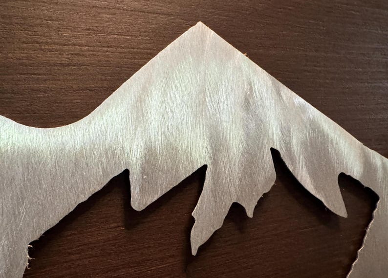 K&ouml;nnte beinhalten: Ein silberfarbener Metallzuschnitt in Form eines Gebirgszuges mit geb&uuml;rsteter Oberfl&auml;che. Das St&uuml;ck weist scharfe, gezackte Gipfel und tiefe T&auml;ler auf und zeigt eine metallische Textur mit sichtbarer, richtungsgebundener Maserung.