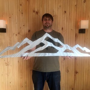 May include: A wide, silver-colored metal wall art piece featuring a stylized mountain range silhouette with jagged peaks and cut-out sections. The surface of the metal has a brushed, linear texture that reflects light across the mountain shapes.