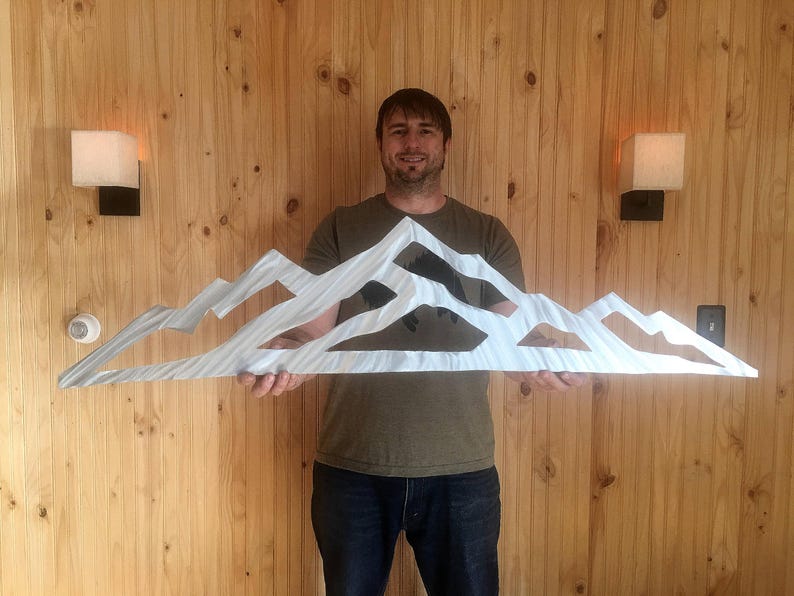 May include: A wide, silver-colored metal wall art piece featuring a stylized mountain range silhouette with jagged peaks and cut-out sections. The surface of the metal has a brushed, linear texture that reflects light across the mountain shapes.