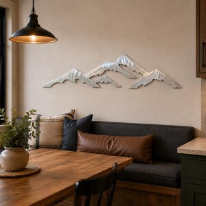 May include: A horizontal silver-toned metal wall art piece depicting a mountain range with jagged, layered peaks. The sculpture features a brushed metallic finish and is mounted on a plain wall above a dining area with a wooden table and bench.
