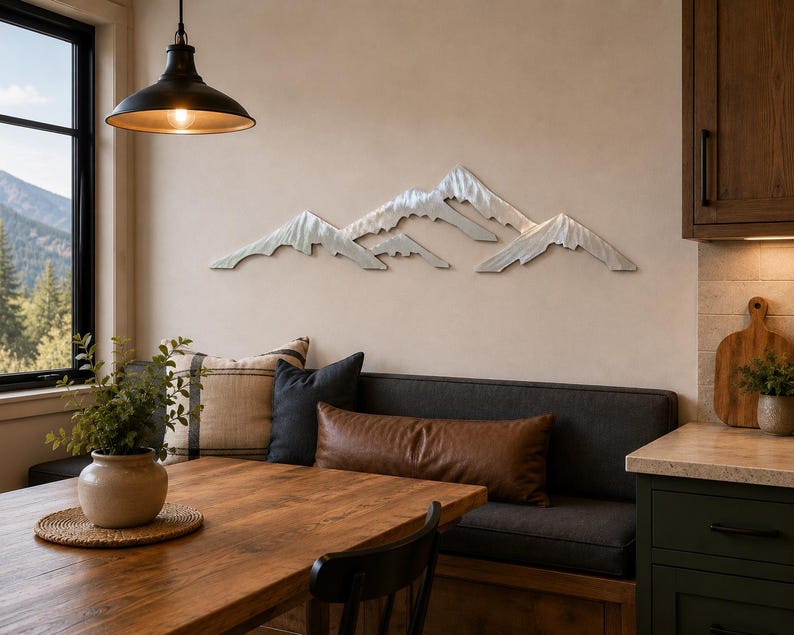 May include: A horizontal silver-toned metal wall art piece depicting a mountain range with jagged, layered peaks. The sculpture features a brushed metallic finish and is mounted on a plain wall above a dining area with a wooden table and bench.