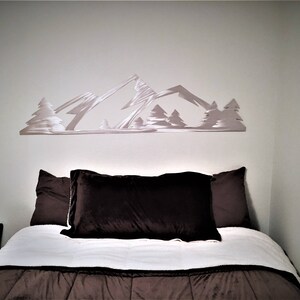 May include: A white metal mountain range wall decor with trees, above a white and brown bed with two brown pillows.