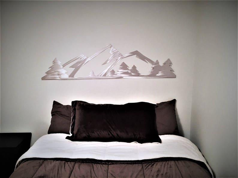 May include: A white metal mountain range wall decor with trees, above a white and brown bed with two brown pillows.