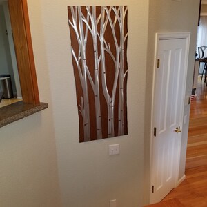 May include: Metal wall art featuring a stylized depiction of birch trees. The artwork has a brown background with silver-colored tree trunks and branches. The piece is rectangular and mounted on a light-colored wall.