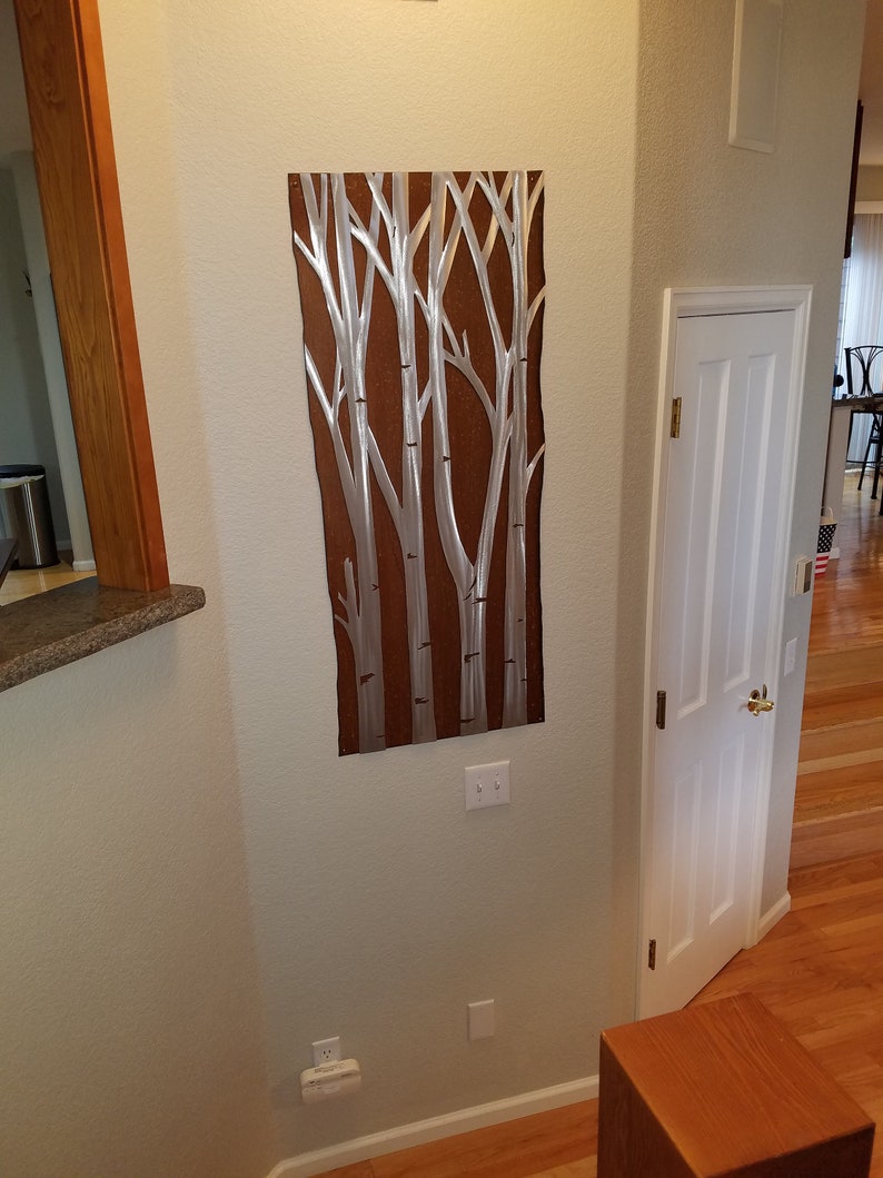 May include: Metal wall art featuring a stylized depiction of birch trees. The artwork has a brown background with silver-colored tree trunks and branches. The piece is rectangular and mounted on a light-colored wall.