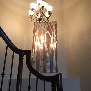 May include: A decorative wall art piece featuring a birch tree design with silver and white tree trunks against a brown background. A chandelier hangs above the artwork. The image is taken from a low angle, showing a staircase.