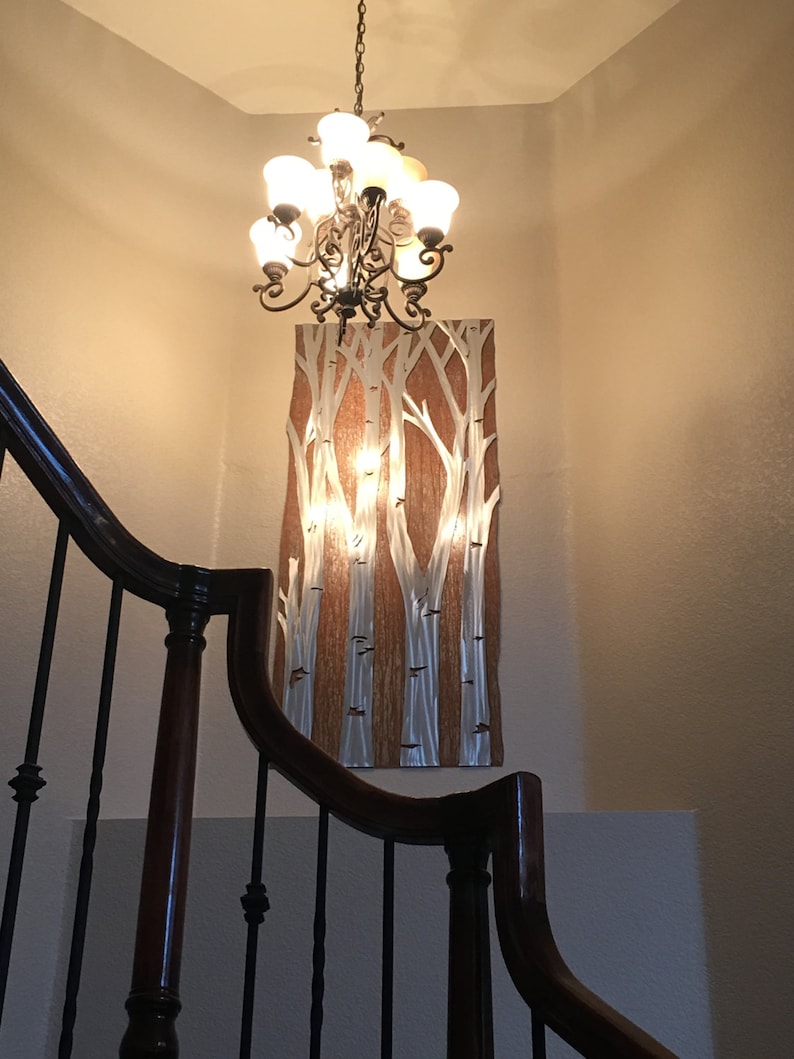 May include: A decorative wall art piece featuring a birch tree design with silver and white tree trunks against a brown background. A chandelier hangs above the artwork. The image is taken from a low angle, showing a staircase.