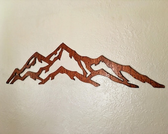 May include: A wall-mounted mountain range silhouette crafted from dark brown wood with a visible grain texture. The design features jagged, angular peaks and valleys, creating a wide, horizontal landscape cutout that displays negative space.