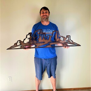 Puede incluir: Una obra de arte mural de madera que representa un paisaje des&eacute;rtico con mesas y buttes. La obra est&aacute; hecha de madera en capas, con tonos de madera natural y acentos blancos. La pieza la sostiene una persona con una camisa azul y pantalones cortos grises.