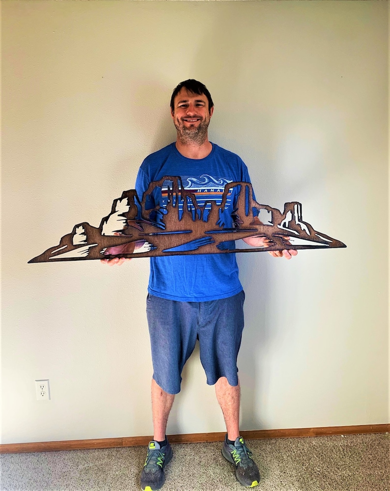 Puede incluir: Una obra de arte mural de madera que representa un paisaje des&eacute;rtico con mesas y buttes. La obra est&aacute; hecha de madera en capas, con tonos de madera natural y acentos blancos. La pieza la sostiene una persona con una camisa azul y pantalones cortos grises.