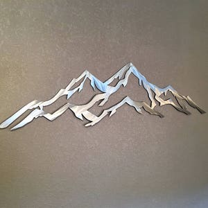 May include: A silver metal wall sculpture depicting a mountain range with three peaks. The sculpture is cut out and has a smooth, reflective surface.