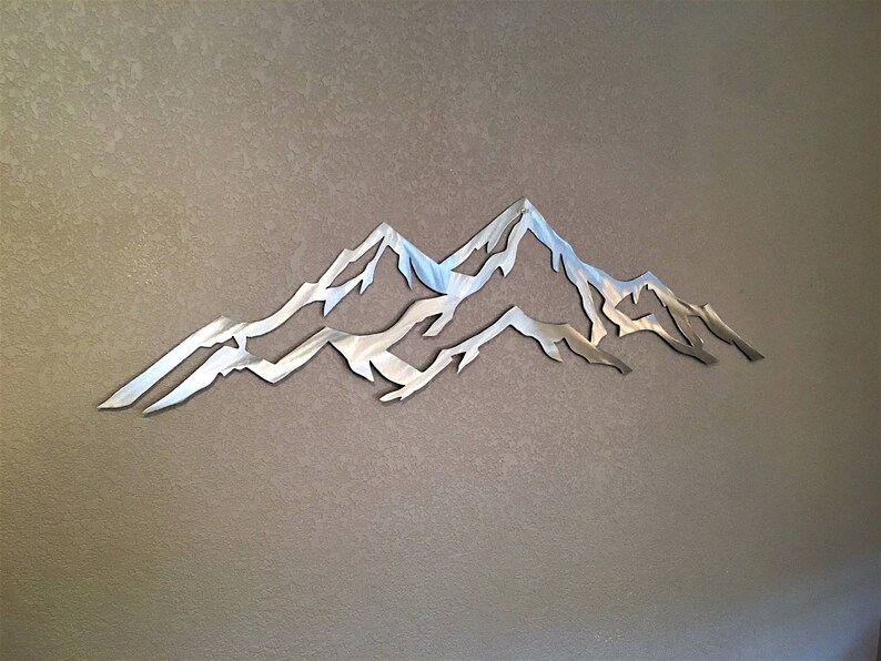 May include: A silver metal wall sculpture depicting a mountain range with three peaks. The sculpture is cut out and has a smooth, reflective surface.