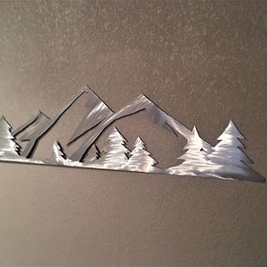 May include: A metal wall art sculpture depicting a mountain range with a silhouette of trees in front. The sculpture is made of silver metal and has a brushed finish.