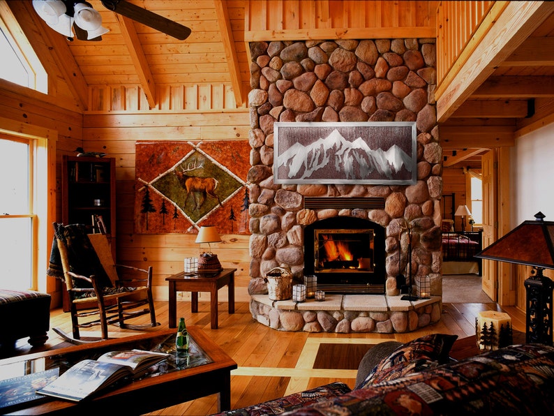 May include: A cozy living room with a stone fireplace, wooden floors, and walls. A painting of a deer hangs on the wall. A metal mountain art piece is above the fireplace. A rocking chair and coffee table complete the rustic decor.