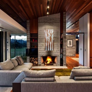 May include: A modern living room with a fireplace, a large gray sectional sofa, and floor-to-ceiling windows. The room features a wooden ceiling and walls, and a painting of white birch trees.
