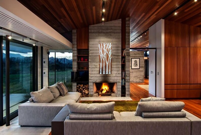 May include: A modern living room with a fireplace, a large gray sectional sofa, and floor-to-ceiling windows. The room features a wooden ceiling and walls, and a painting of white birch trees.