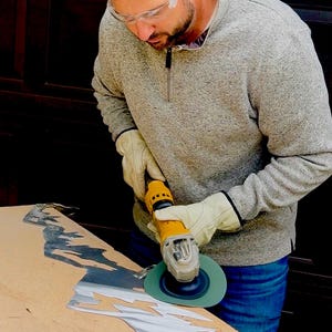 May include: A yellow power tool with a green circular sanding disc is being used to smooth the edges of a jagged, silver-colored metal sheet. The tool is held by hands wearing beige leather work gloves, while clear safety glasses are worn.