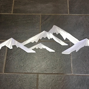 May include: A set of four white metal mountain silhouettes with pointed peaks and jagged edges. The pieces are arranged in a horizontal line, featuring varying heights and widths to create a layered, multi-peak mountain range effect.