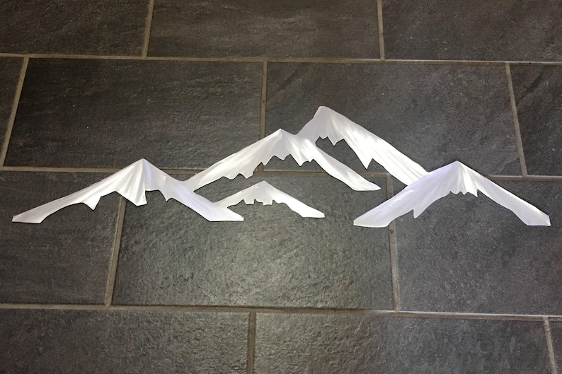 May include: A set of four white metal mountain silhouettes with pointed peaks and jagged edges. The pieces are arranged in a horizontal line, featuring varying heights and widths to create a layered, multi-peak mountain range effect.