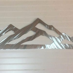 May include: A silver-colored metal wall art piece featuring a stylized mountain range silhouette. The design consists of multiple overlapping peaks with a brushed, reflective finish that creates a textured appearance across the surface.