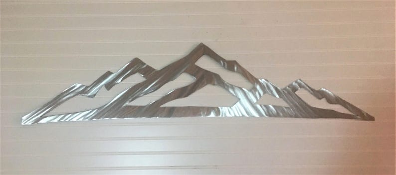 May include: A silver-colored metal wall art piece featuring a stylized mountain range silhouette. The design consists of multiple overlapping peaks with a brushed, reflective finish that creates a textured appearance across the surface.