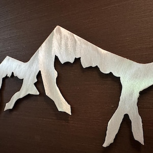 K&ouml;nnte beinhalten: Ein silberfarbenes Wandkunstobjekt aus Metall, das die Silhouette eines zerkl&uuml;fteten Gebirgszuges darstellt. Die Metalloberfl&auml;che weist ein geb&uuml;rstetes, strukturiertes Finish mit sichtbarer Maserung auf, das f&uuml;r einen Glanzeffekt sorgt.