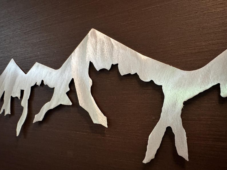 K&ouml;nnte beinhalten: Ein silberfarbenes Wandkunstobjekt aus Metall, das die Silhouette eines zerkl&uuml;fteten Gebirgszuges darstellt. Die Metalloberfl&auml;che weist ein geb&uuml;rstetes, strukturiertes Finish mit sichtbarer Maserung auf, das f&uuml;r einen Glanzeffekt sorgt.