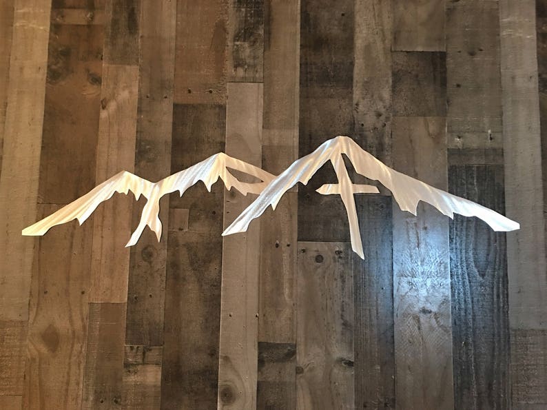 May include: A metal wall decor featuring a silhouette of three mountain peaks. The peaks are cut out of a single sheet of metal and have a brushed silver finish.
