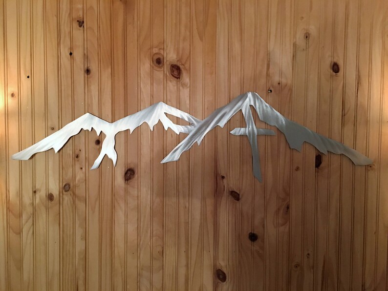 May include: A silver metal mountain range wall art sculpture with a jagged, pointed peak. The sculpture is mounted on a wood panel wall.