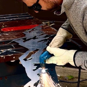 May include: A person wearing protective eyewear and beige leather gloves uses a blue plasma cutter to carve a jagged mountain range design into a flat sheet of dark gray metal, creating bright orange sparks at the point of contact.