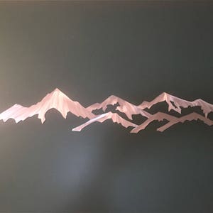 May include: A metallic wall art piece featuring a stylized mountain range silhouette. The design consists of layered, jagged peaks with a brushed silver finish that reflects light, creating a textured appearance across the horizontal metal structure.