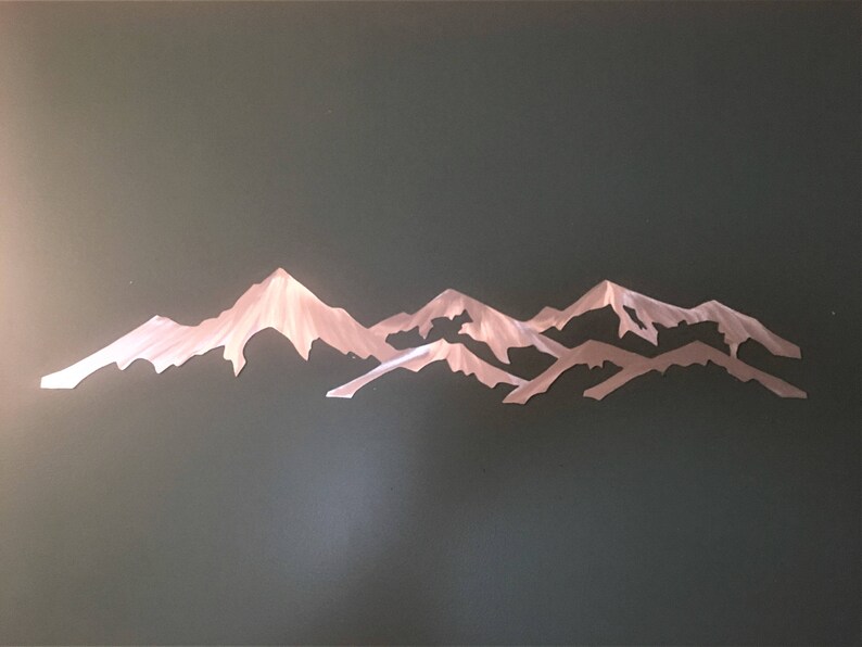 May include: A metallic wall art piece featuring a stylized mountain range silhouette. The design consists of layered, jagged peaks with a brushed silver finish that reflects light, creating a textured appearance across the horizontal metal structure.