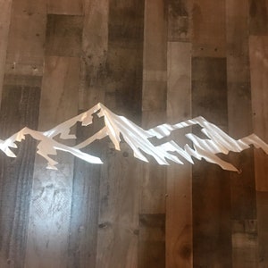 May include: A silver metal mountain range wall art sculpture. The sculpture is made of thin metal and has a brushed finish. The mountains are in a horizontal line and are all different sizes. The sculpture is perfect for adding a modern touch to any room.